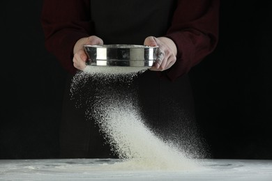 Photo of Woman sieving flour at table against black background, closeup