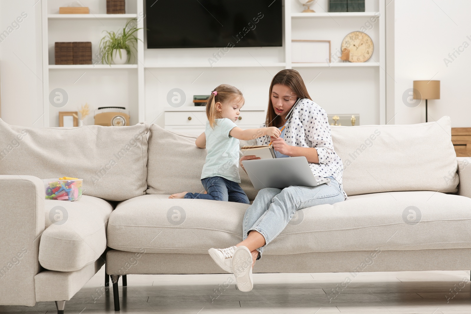 Photo of Woman working remotely at home. Little daughter bothering her mother on sofa in living room