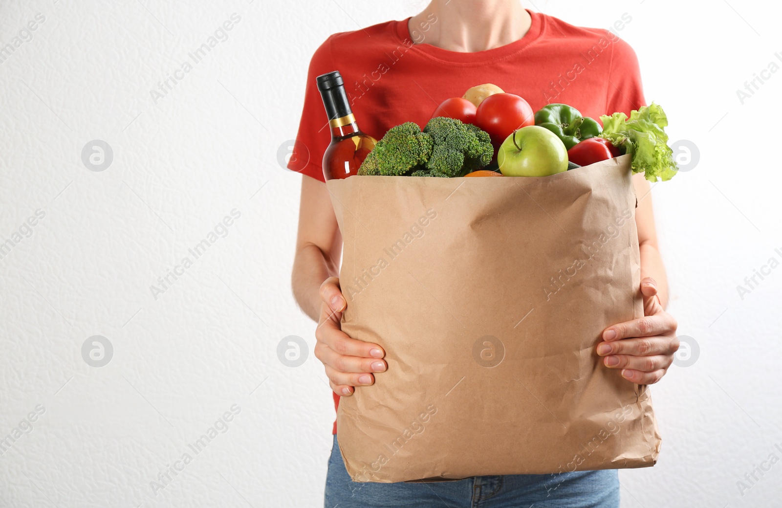 Photo of Woman holding paper bag with different groceries near white wall, closeup view. Space for text