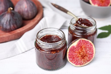 Photo of Glass jars of tasty sweet fig jam with spoon and fruits on white wooden table