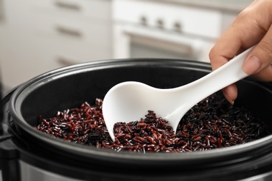 Photo of Woman taking brown rice from multi cooker with spoon in kitchen, closeup