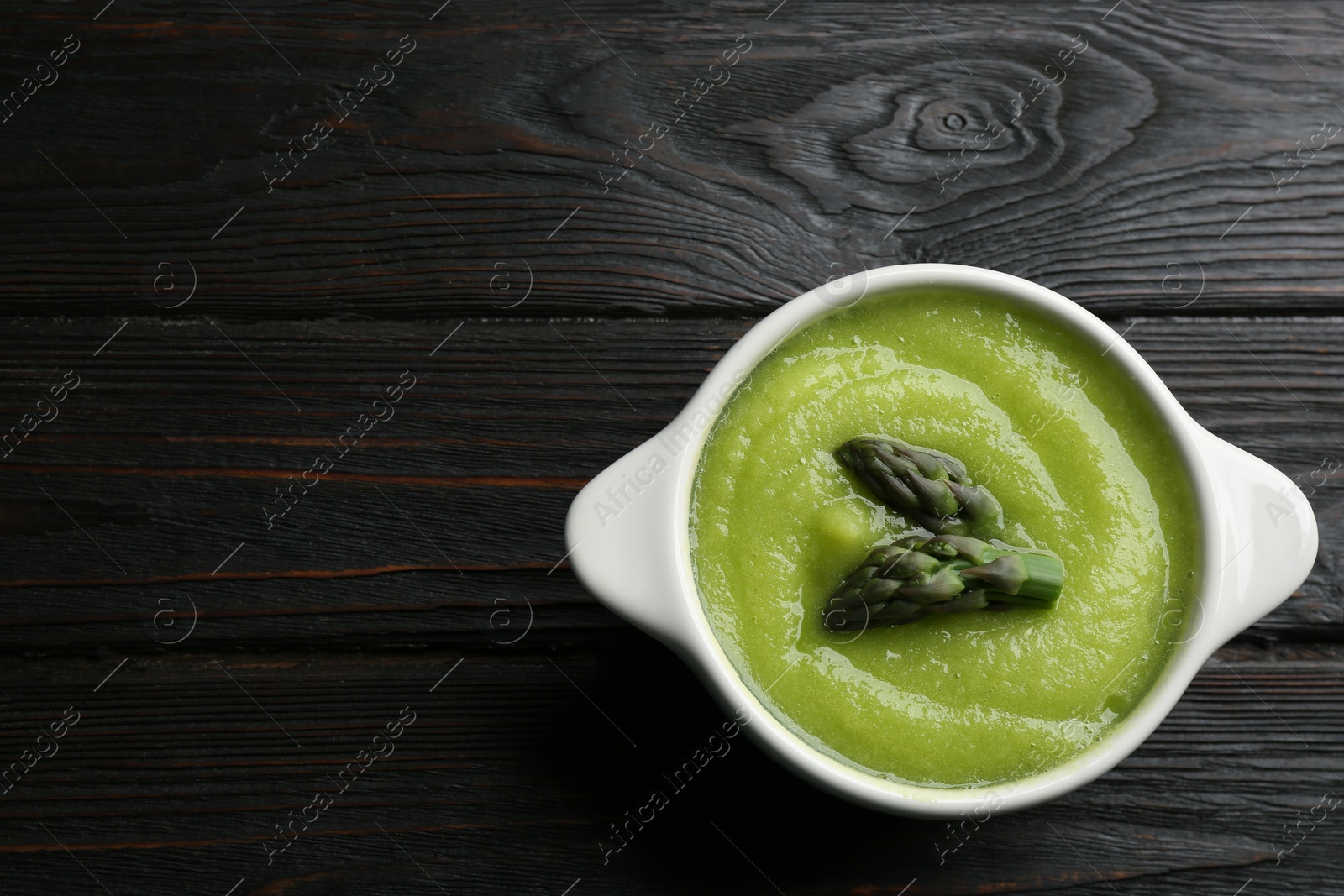 Photo of Delicious asparagus soup in bowl on black wooden table, top view. Space for text