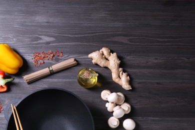 Photo of Empty iron wok, chopsticks and ingredients on dark grey wooden table, flat lay. Space for text