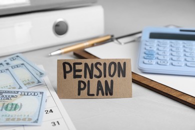 Photo of Card with phrase Pension Plan, calculator, dollar banknotes and calendar on white office table, closeup