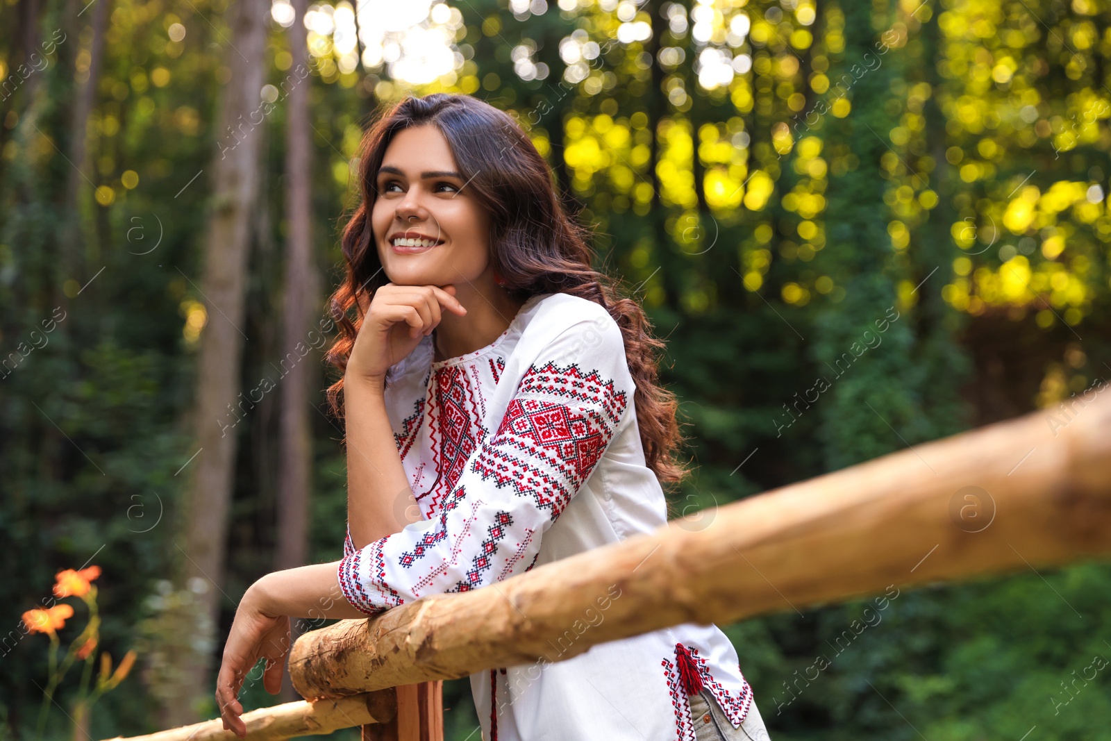 Photo of Beautiful woman wearing embroidered shirt near wooden railing in countryside. Ukrainian national clothes