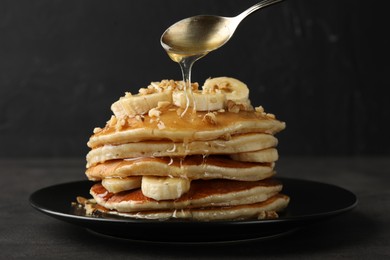 Photo of Pouring honey from spoon onto delicious pancakes with bananas and walnuts at dark table, closeup