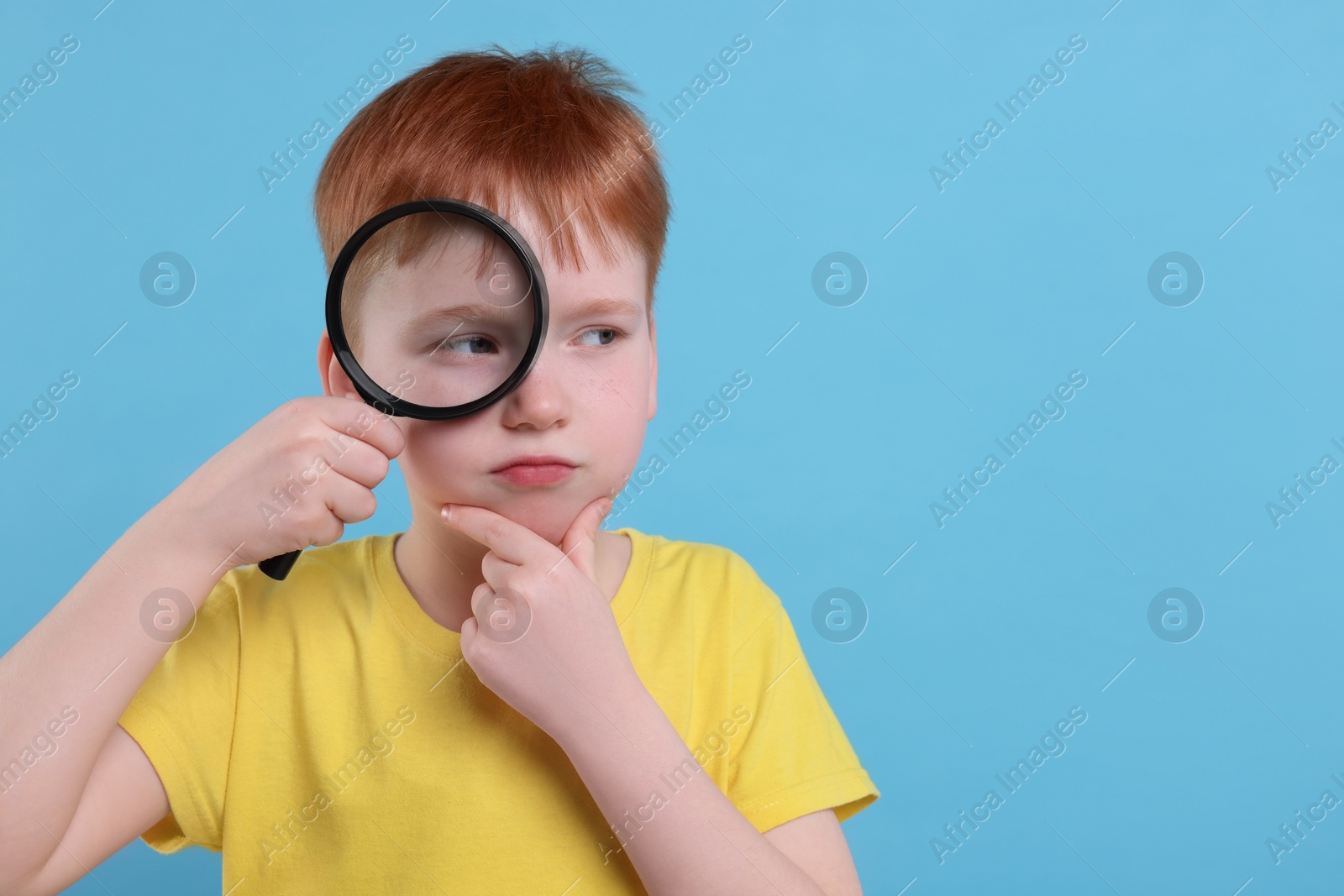 Photo of Thoughtful boy looking through magnifier glass on light blue background, space for text