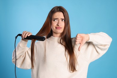 Photo of Upset young woman with flattening iron showing thumb down on light blue background. Hair damage