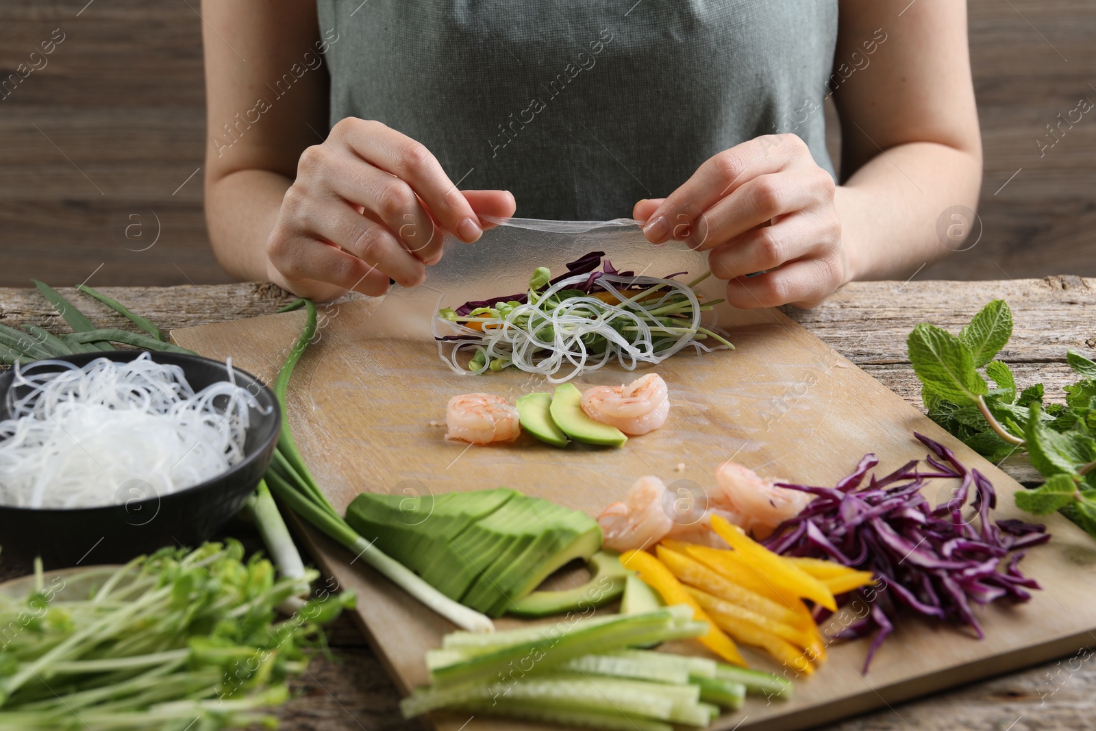 Photo of Woman wrapping spring roll at wooden table with products, closeup