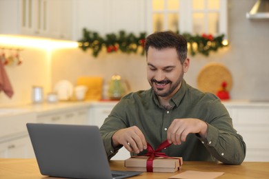 Photo of Celebrating Christmas online with exchanged by mail presents. Happy man opening gift box during video call on laptop at home