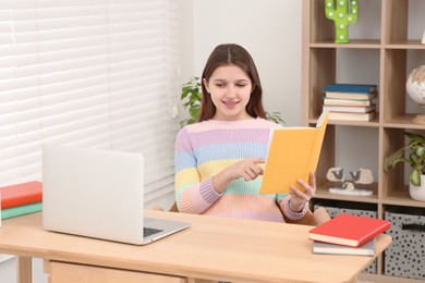 Photo of Cute girl reading book near laptop at desk in room. Home workplace