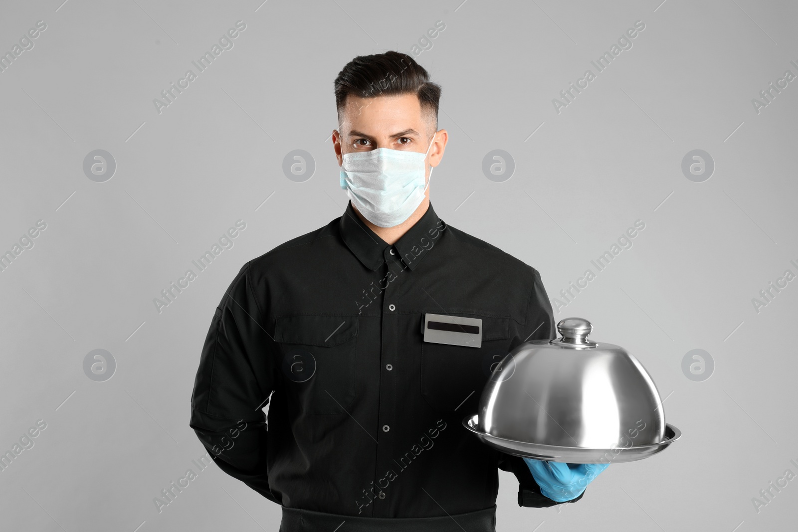 Photo of Waiter in medical face mask holding tray with lid on light grey background