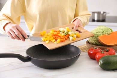 Photo of Woman pouring mix of cut vegetables into frying pan at table in kitchen, closeup