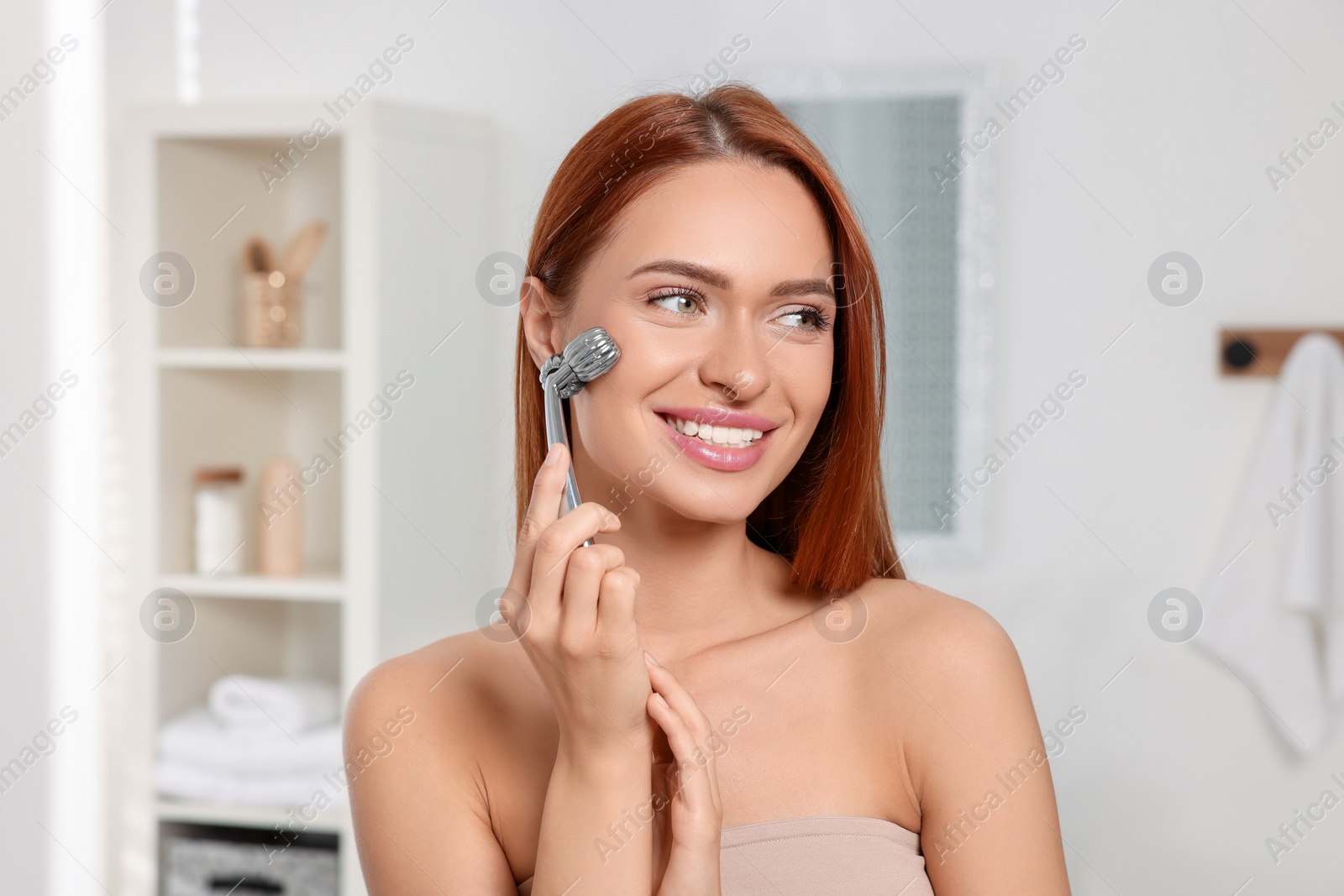 Photo of Young woman massaging her face with metal roller in bathroom