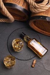 Photo of Whiskey with ice cubes in glasses, bottle, wooden barrels and rope on black table, flat lay