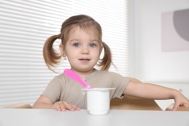 Cute little child eating tasty yogurt from plastic cup with spoon at white table indoors