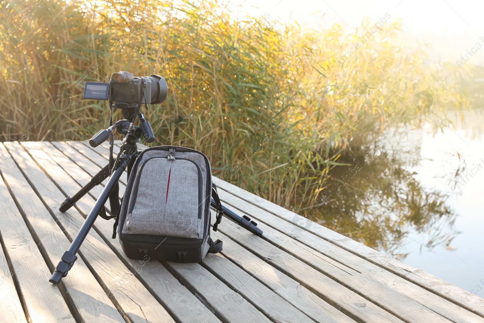 Photo of Tripod with modern camera and backpack on wooden pier near water. Professional photography