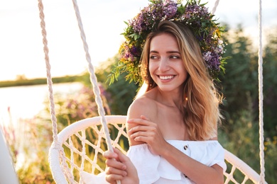Photo of Young woman wearing wreath made of beautiful flowers on swing chair outdoors