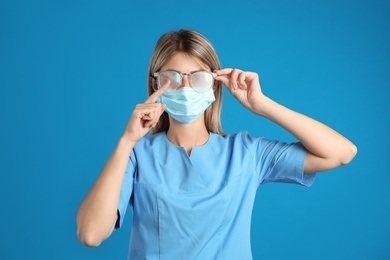 Photo of Doctor wiping foggy glasses caused by wearing disposable mask on blue background. Protective measure during coronavirus pandemic