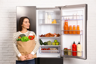 Young woman with paper bag full of products near open refrigerator indoors