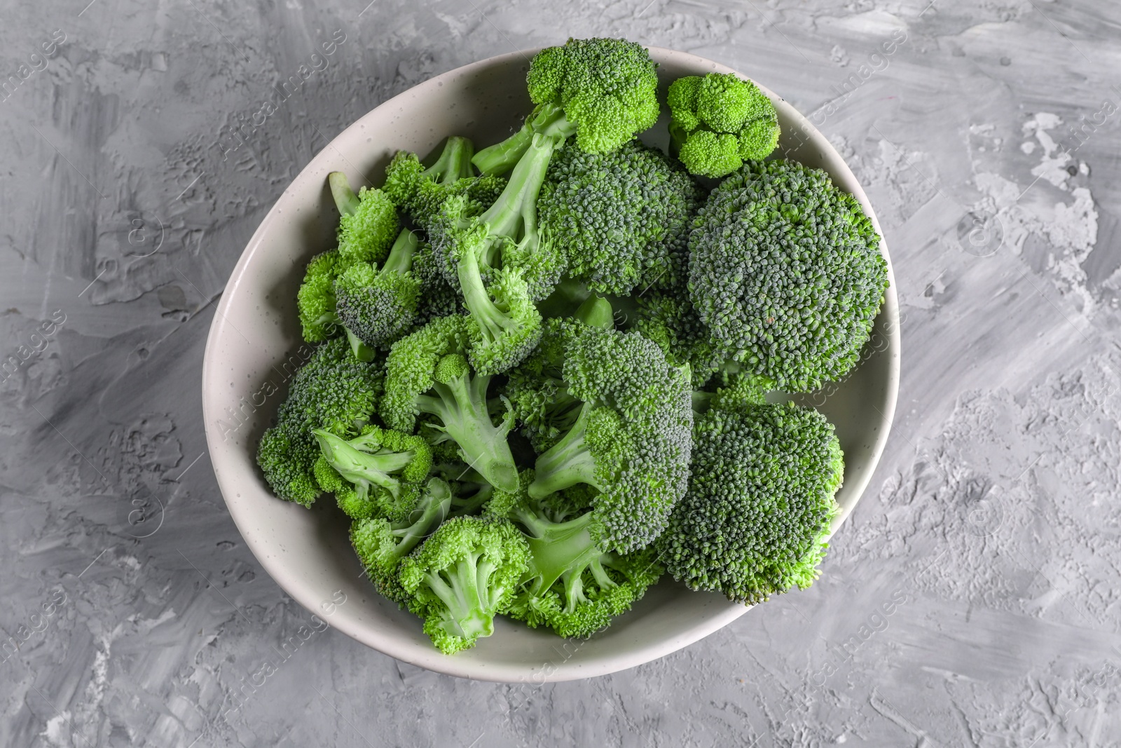 Photo of Bowl of fresh raw broccoli on grey textured table, top view