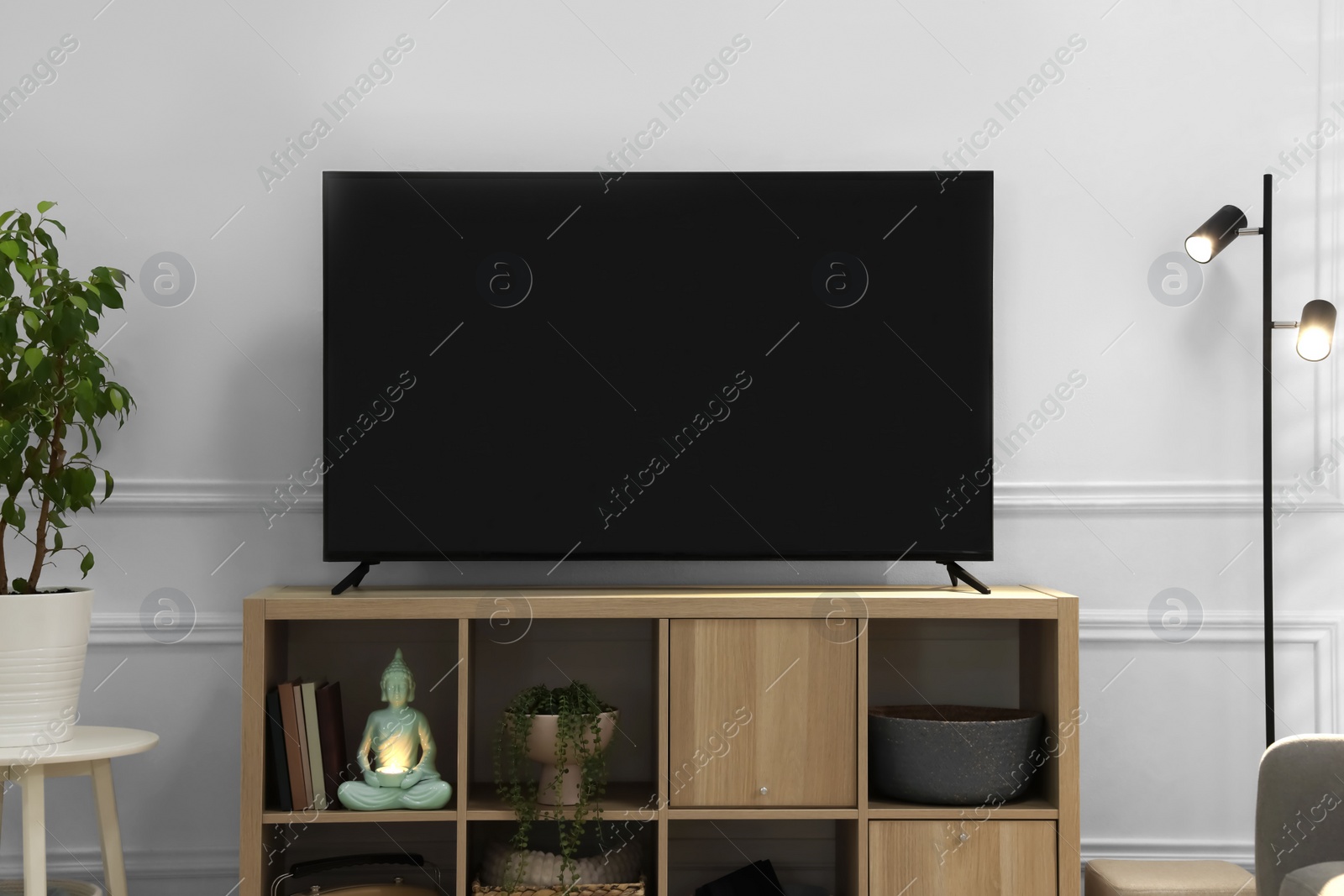 Photo of Modern TV on cabinet, lamp and beautiful houseplants near white wall indoors. Interior design