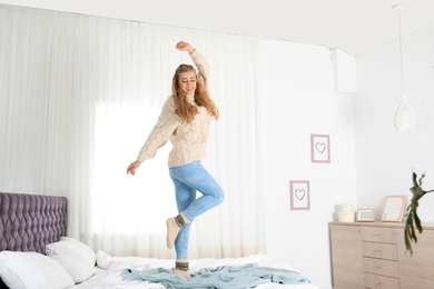 Photo of Attractive smiling young woman in cozy warm sweater jumping on bed at home