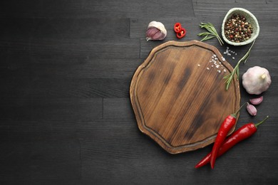 Photo of Cutting board, spices, garlic, rosemary and chili peppers on black wooden table, flat lay. Space for text