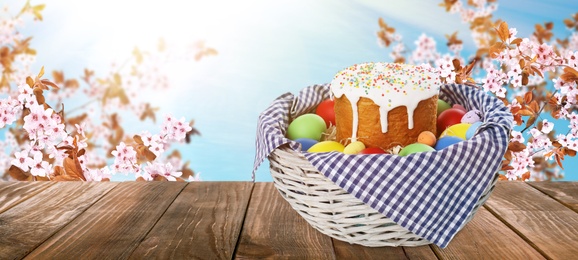 Basket with traditional Easter cake and eggs on wooden table outdoors, space for text. Banner design