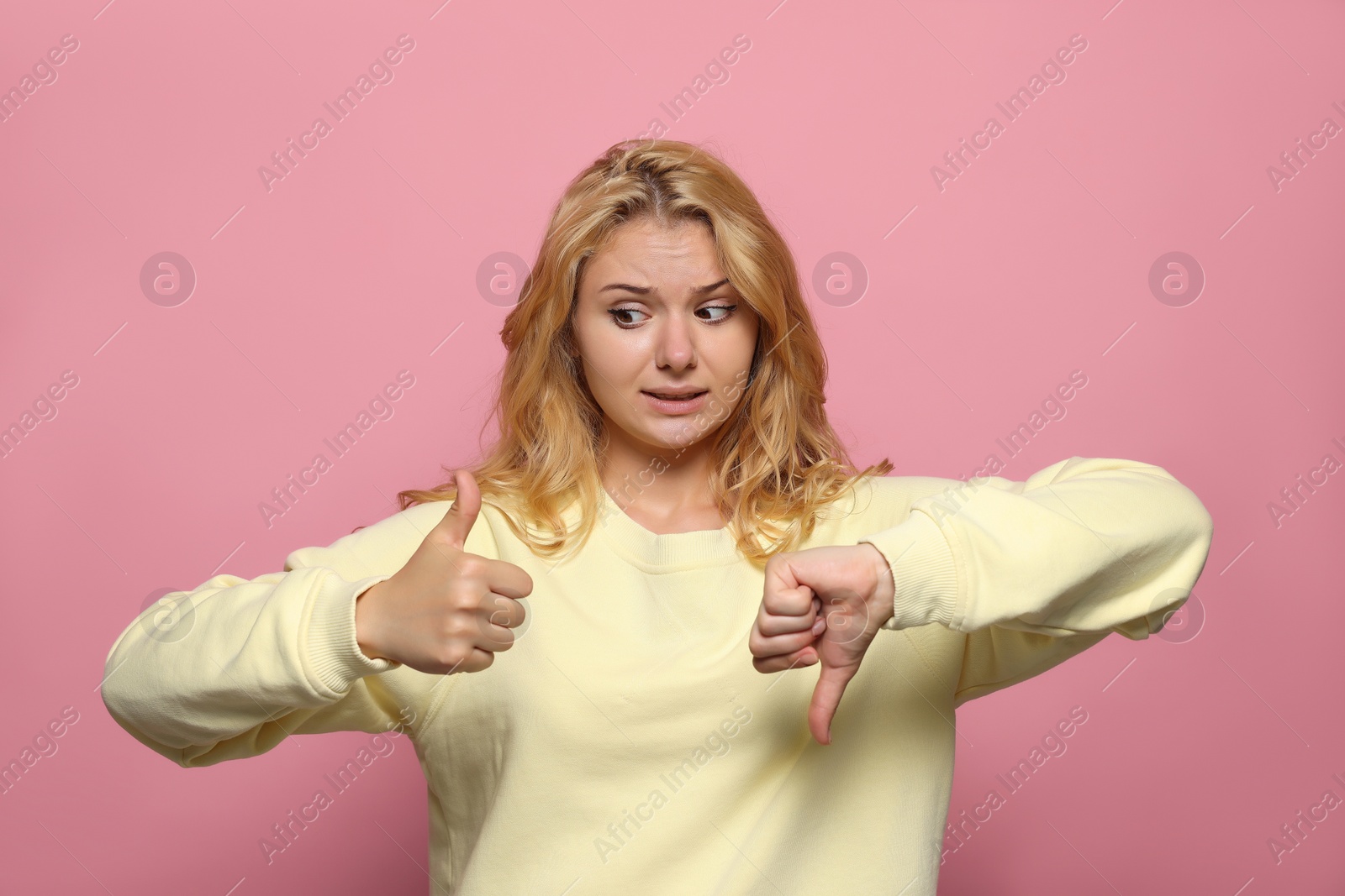 Photo of Conflicted young woman showing thumbs up and down gestures on pink background