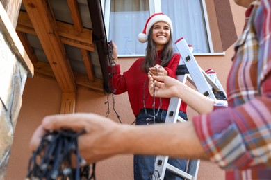 Photo of Happy woman in Santa hat and man decorating house with Christmas lights outdoors, low angle view