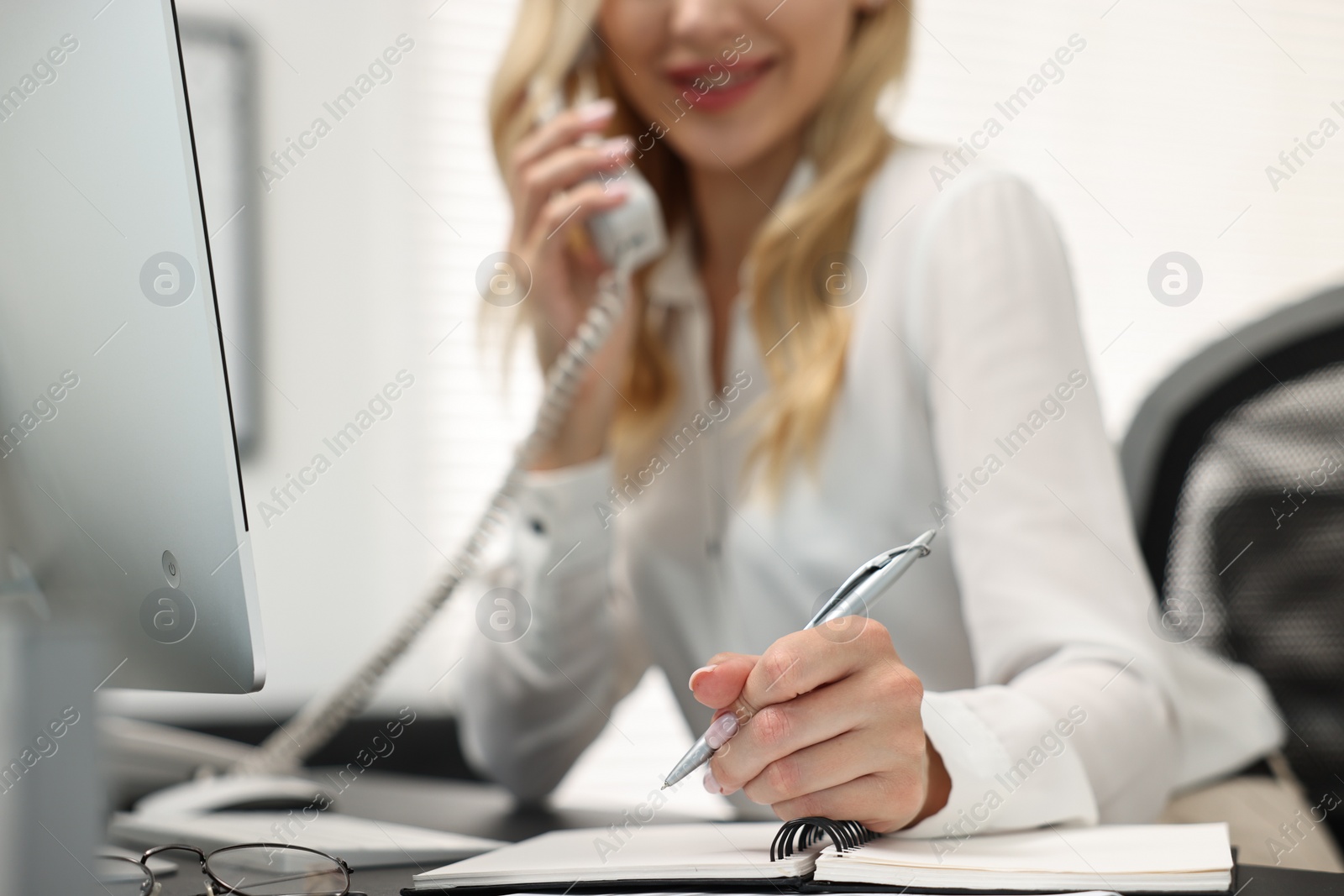 Photo of Secretary talking on phone and taking notes at table in office, closeup