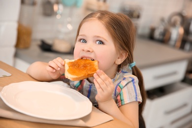 Photo of Little girl eating toast with jam at table in kitchen