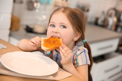 Little girl eating toast with jam at table in kitchen