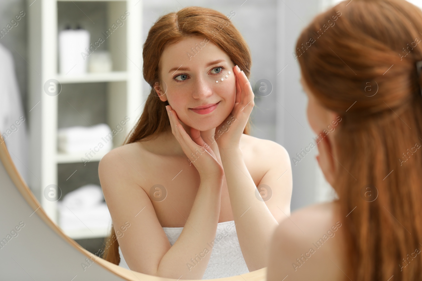 Photo of Beautiful woman with freckles and cream on her face near mirror in bathroom