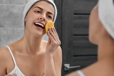 Young woman with headband washing her face using sponge near mirror in bathroom