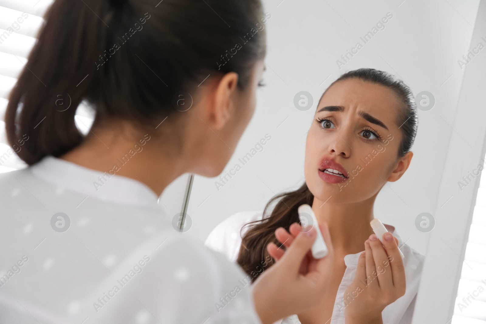 Photo of Young woman with herpes applying lip balm in front of mirror at home