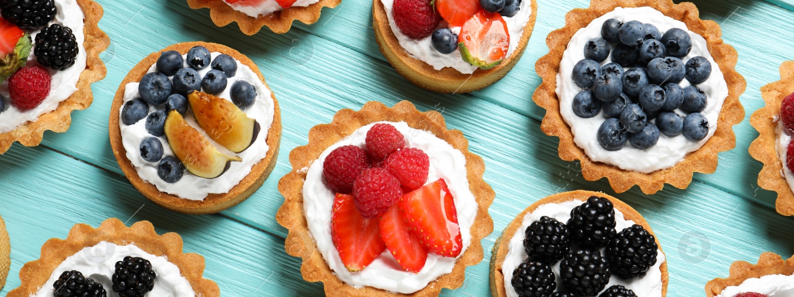 Image of Many different tasty tartlets with fresh berries on light blue wooden table, flat lay. Banner design