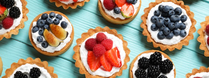 Many different tasty tartlets with fresh berries on light blue wooden table, flat lay. Banner design