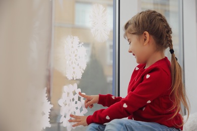 Photo of Cute little girl decorating window with paper snowflake indoors