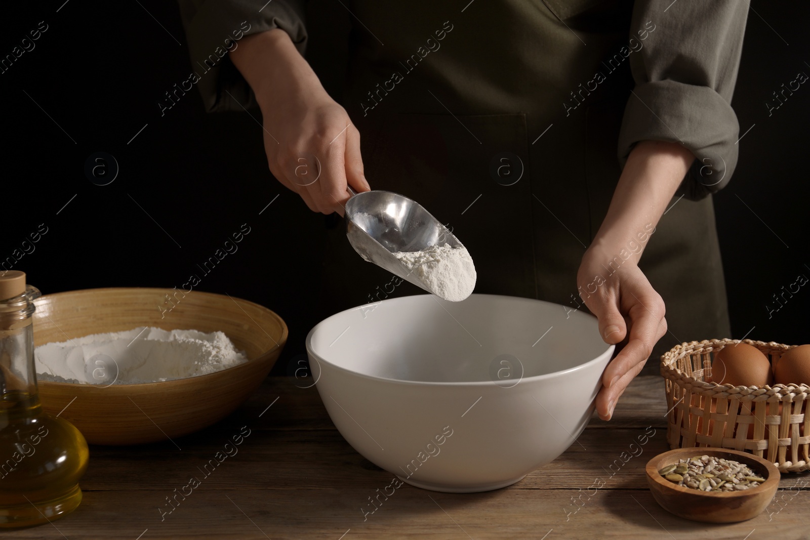 Photo of Making bread. Woman adding flour into bowl at wooden table on dark background, closeup