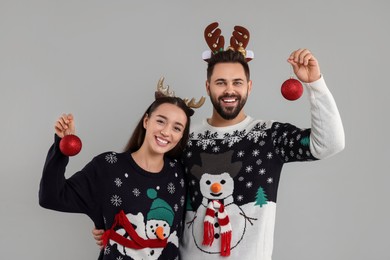 Photo of Happy young couple in Christmas sweaters and reindeer headbands holding festive baubles on grey background