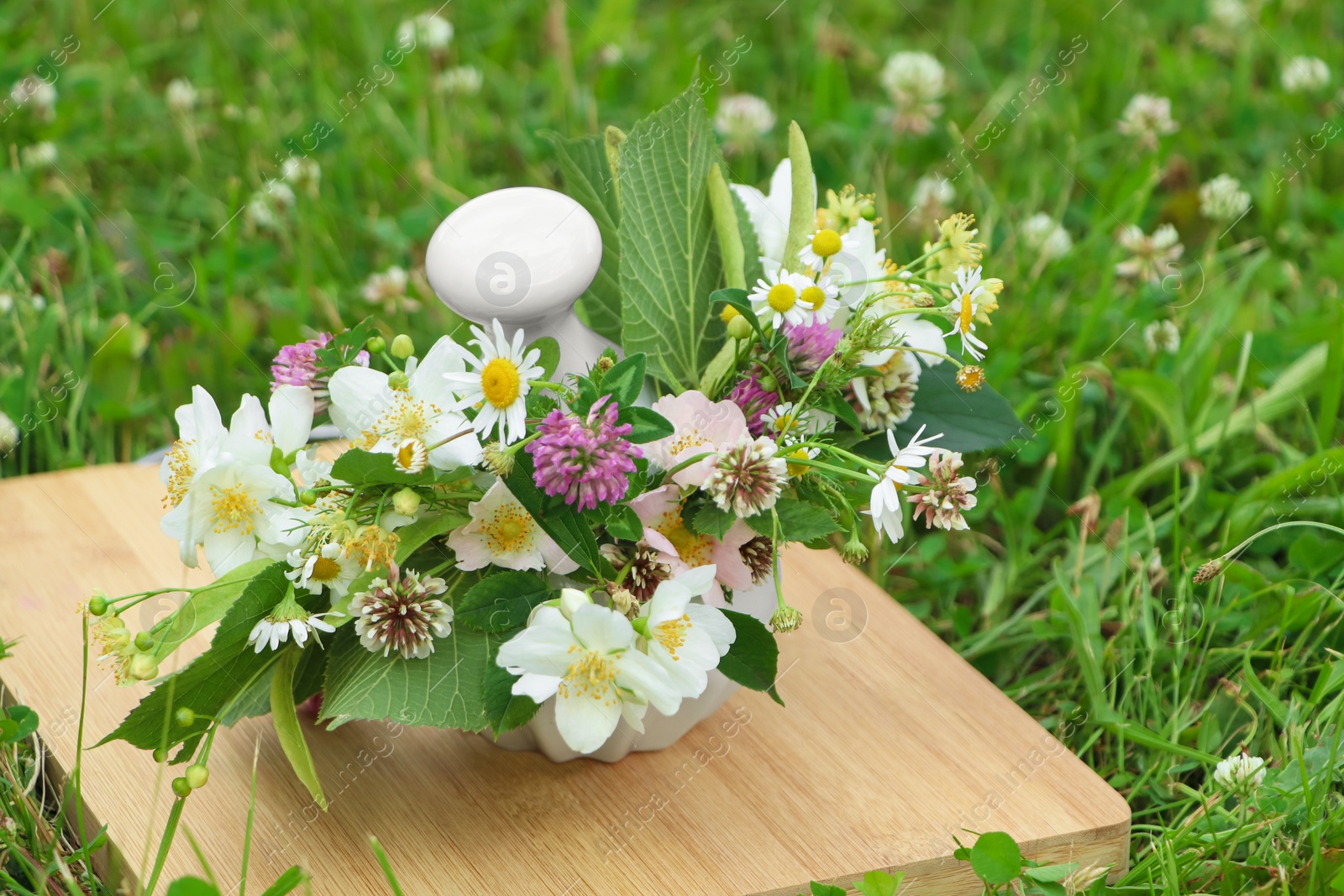 Photo of Ceramic mortar with pestle, different wildflowers and herbs on wooden board in meadow