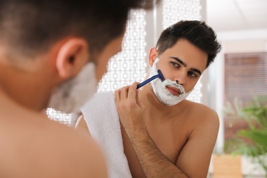 Photo of Handsome young man shaving with razor near mirror in bathroom