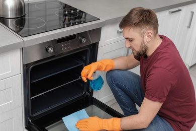 Photo of Young man cleaning oven with rag and detergent in kitchen