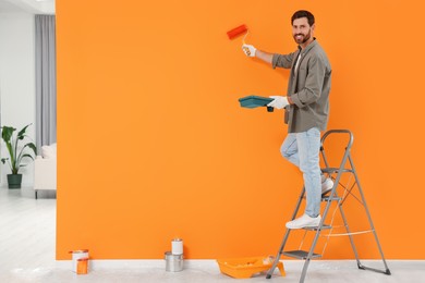 Photo of Happy designer painting orange wall with roller indoors, space for text