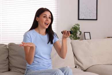 Emotional woman with remote controller watching TV on sofa at home