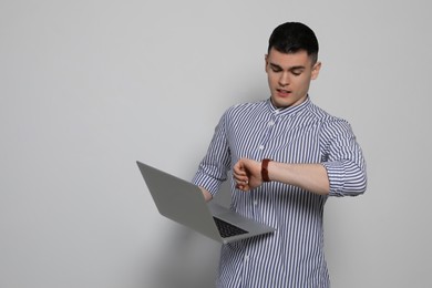 Emotional young man with laptop checking time on light grey background, space for text. Being late concept