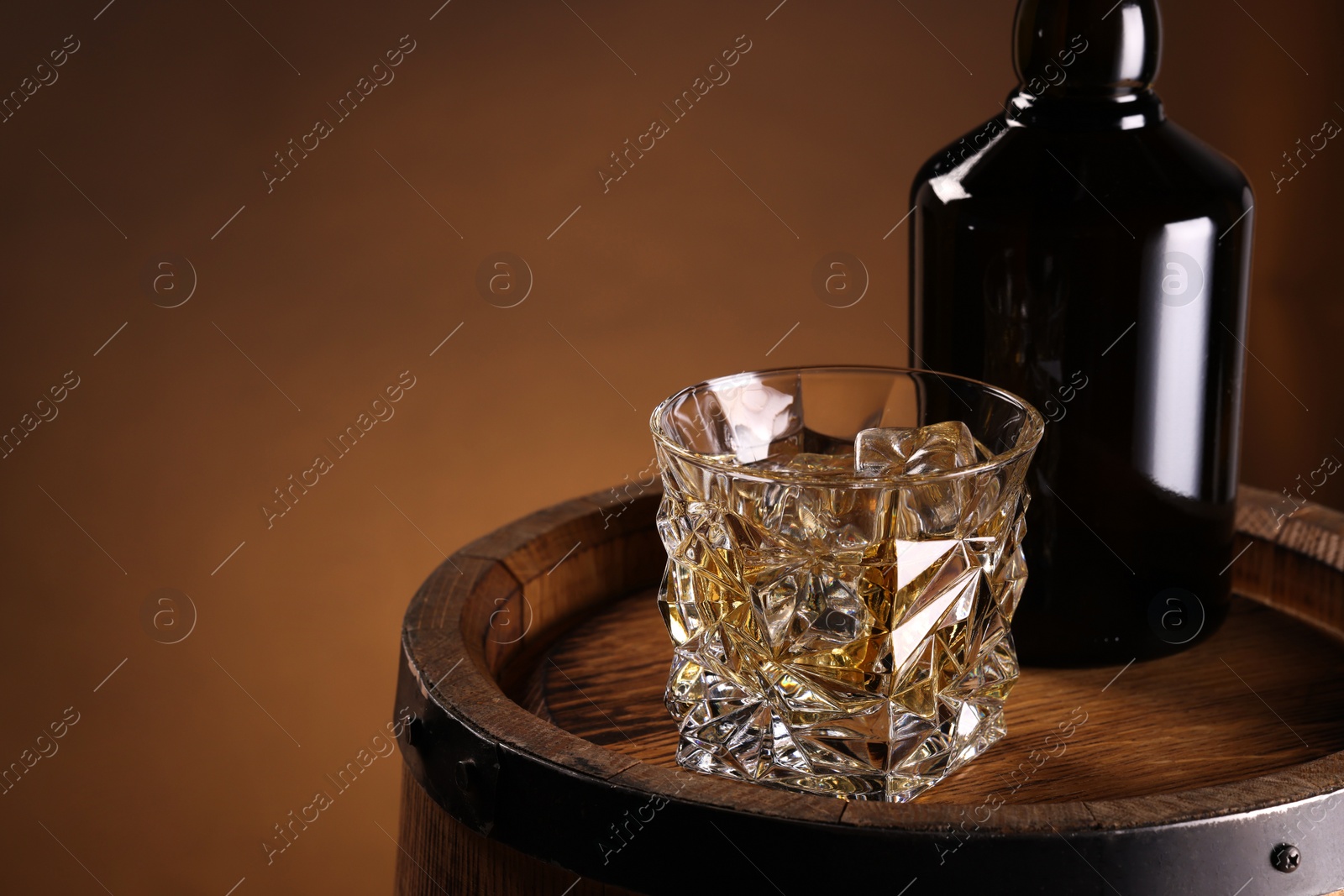 Photo of Whiskey with ice cubes in glass and bottle on wooden barrel against brown background, space for text