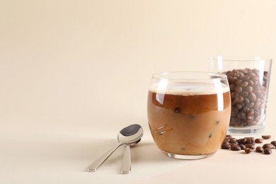 Photo of Refreshing iced coffee with milk in glass, beans and spoons on beige background, space for text